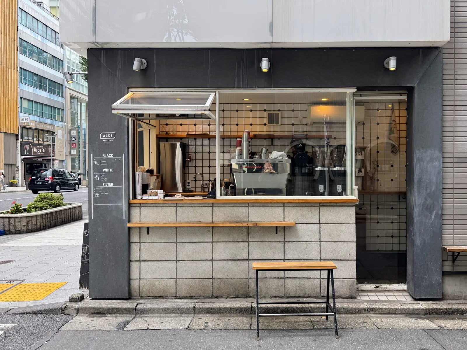 The street counter at About Life Coffee Brewers in Shibuya: a serving hatch and a window onto an espresso machine and two grinders, a short menu in three sections, and a single bench on the pavement.